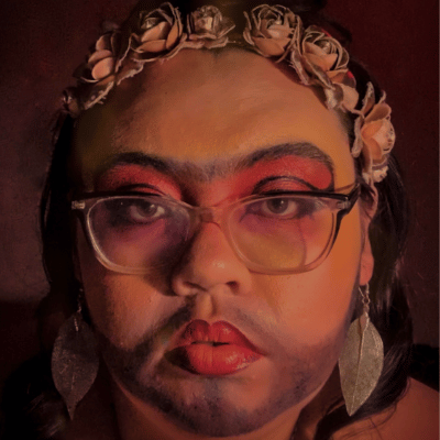 A portrait of Jazz. Jazz has light skin and dark hair in a bun on the top of their head with two strands of hair framing their face. They are wearing glasses, a rose gold flower crown, a red floral hairpin, and silver leaf earrings. They have on bright red eyeshadow, yellow paint on their face, and their lipstick fades from yellow in the center to dark red.
